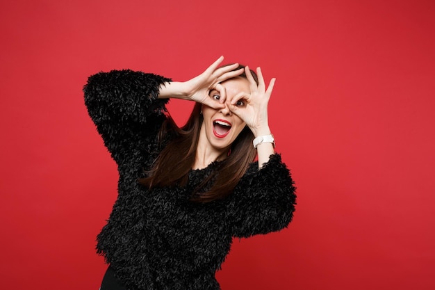 Cheerful young woman in black fur sweater holding hands near eyes imitating glasses or binoculars isolated on bright red wall background. People sincere emotions lifestyle concept. Mock up copy space.