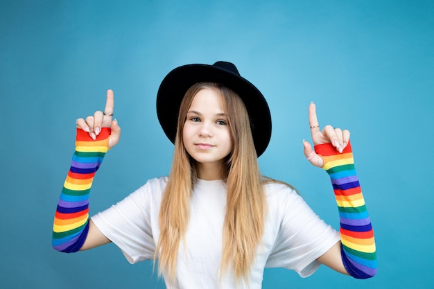 Cheerful young woman in black hat white tshirt and colored\
sleeves pointing with fingers to the side at copy space showing\
cool advertisement on blue background