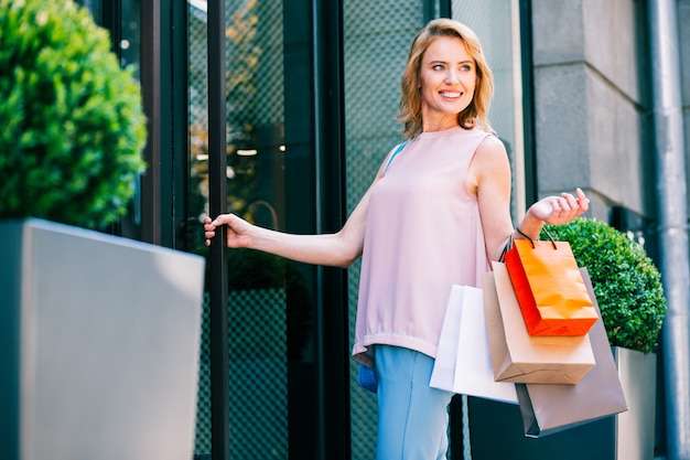 幸せそうに見える買い物袋とガラスのドアのそばの陽気な若い女性