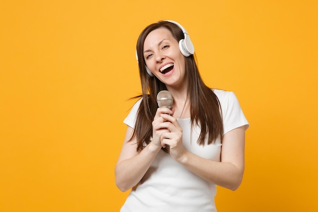 Cheerful young woman in casual clothes with headphones looking\
camera, sing song in microphone spreading hands isolated on yellow\
orange wall background. people lifestyle concept. mock up copy\
space.