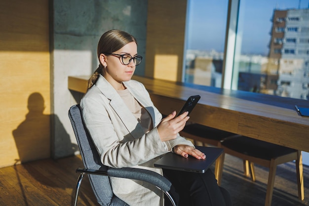 Cheerful young woman in casual wear having telephone conversation sitting at working place near laptop in office smiling female freelancer calling to colleague for share good news about business