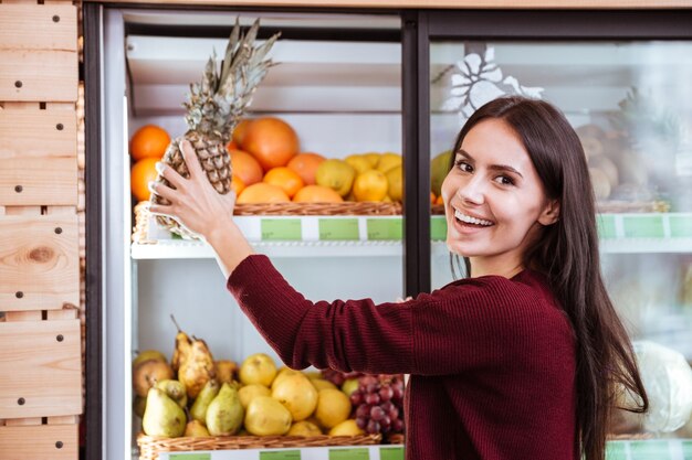 Cheerful young woman choosing and buying pineapple in grocery shop