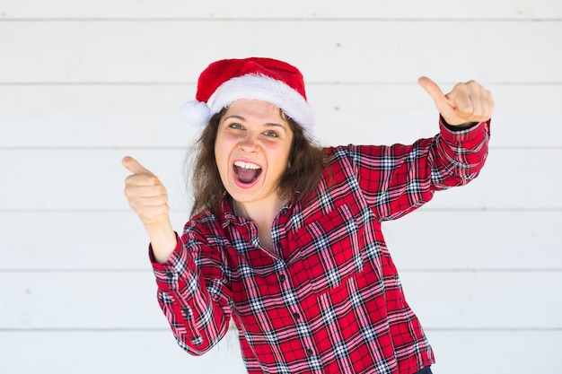 白いスペースに親指を示すクリスマス帽子の陽気な若い女性