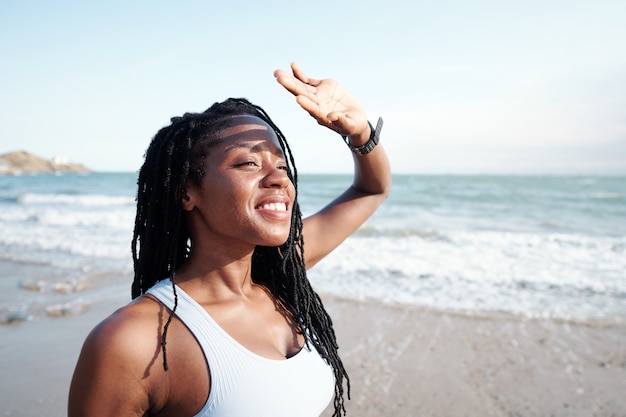 Cheerful young woman covering her eyes with hands from bright sunlight when looking at seascape