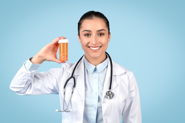 Photo cheerful young woman doctor in uniform showing jar of pills and smiling recommending treatment blue