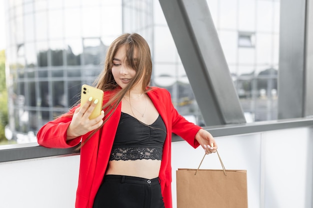 Cheerful young woman dressed in stylish suit holding mobile phone in hand outdoors Beautiful girl holding mobile and standing on street with paper bags