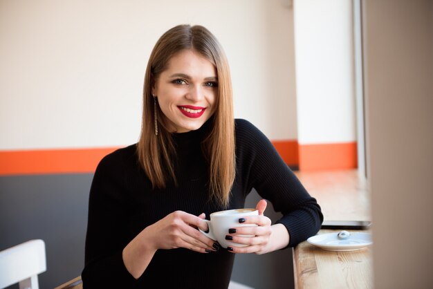 Cheerful young woman drinking warm coffee enjoying it while sitting in cafe. 