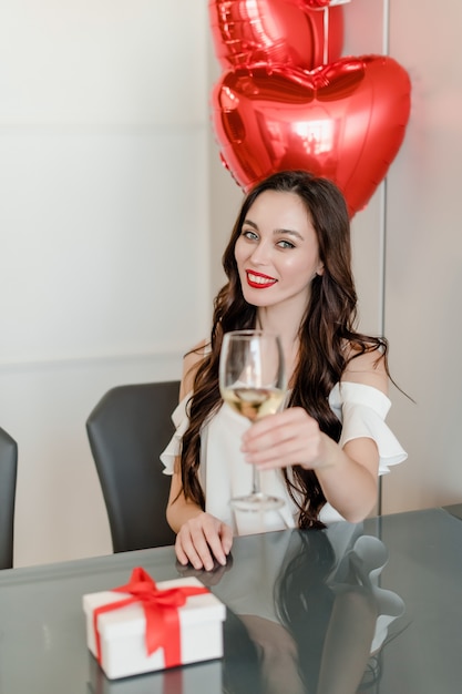 Cheerful young woman drinking wine from glass at home with heart shaped balloons