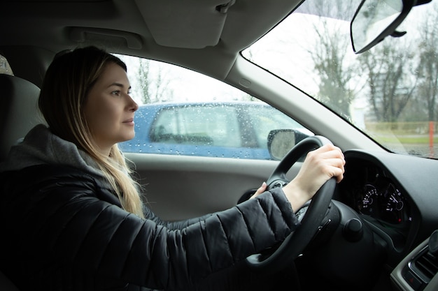 Cheerful young woman driving car, photo inside automobile. Rainy day, lifestyle