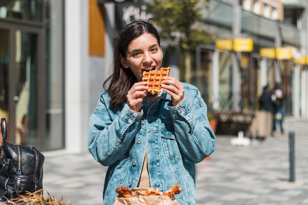 Cheerful young woman enjoying tasty waffles in front of a cafe in the street. take away food in the city