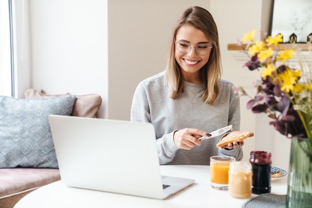 cheerful young woman in eyeglasses eating toasts while using laptop at living room