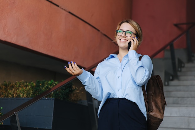 屋外に立っている間モバイル会話を楽しんでいる眼鏡の陽気な若い女性。現代のスマートフォンを使用してカジュアルな服装で幸せなブロンド。