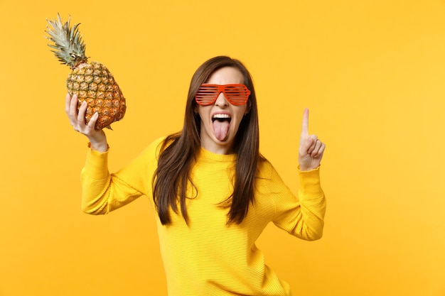 Cheerful young woman in funny glasses showing tongue, pointing index finger up, holding fresh ripe pineapple fruit isolated on yellow orange background. People vivid lifestyle, relax vacation concep