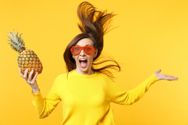 Cheerful young woman in funny glasses with fluttering hair jumping, hold fresh pineapple fruit isolated on yellow orange background. people vivid lifestyle, relax vacation concept. mock up copy spac