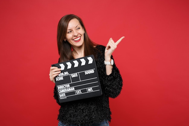 Cheerful young woman in fur sweater pointing index finger aside, hold classic black film making clapperboard isolated on red background. People sincere emotions, lifestyle concept. Mock up copy space.