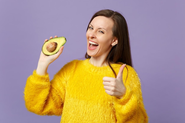 Cheerful young woman in fur sweater showing thumb up, holding half of fresh ripe green avocado isolated on violet pastel background. People vivid lifestyle, relax vacation concept. Mock up copy space.