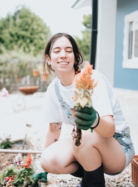 Cheerful young woman in gloves using gardening tools for planting flowers on back yardin casual