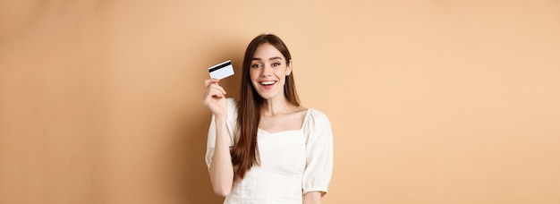 陽気な若い女性は彼女のプラスチック製のクレジット カードを手に入れ、満足して立っているベージュ バックに満足して笑顔