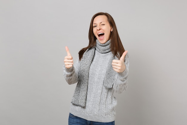 Cheerful young woman in gray sweater, scarf showing thumbs up, blinking isolated on grey wall background. Healthy fashion lifestyle, people sincere emotions, cold season concept. Mock up copy space.