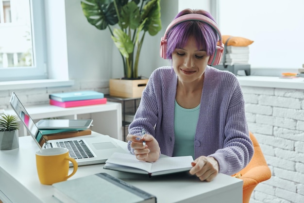 Giovane donna allegra in cuffie che prende appunti mentre si siede al suo posto di lavoro