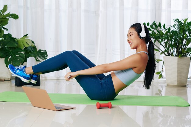 Photo cheerful young woman in headphones repeating after online trainer when doing abs exercise