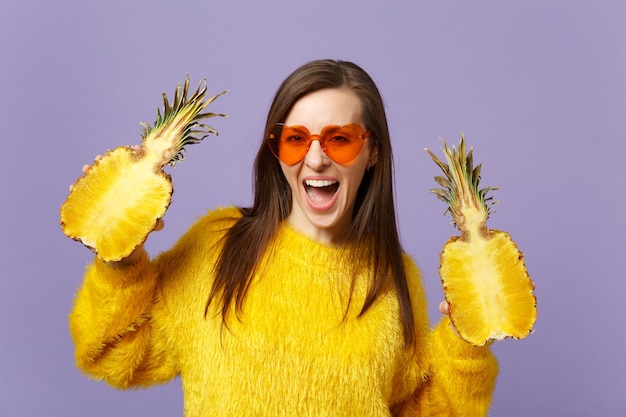 Cheerful young woman in heart glasses keeping mouth open holding halfs of fresh pineapple fruit isolated on violet pastel background. People vivid lifestyle relax vacation concept. Mock up copy space.