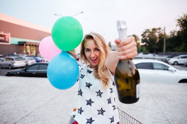 Фото Веселая молодая женщина, держащая бутылку шампанского и воздушные шары на открытом воздухе