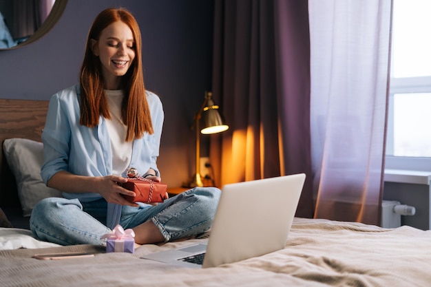 Веселая молодая женщина, держащая подарочную коробку с подарком во время онлайн-общения с помощью ноутбука