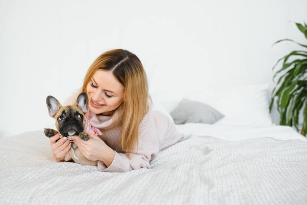 黒い鼻で彼女の大きな子犬を保持し、笑っている陽気な若い女性。フレンチブルドッグとポーズをとって笑顔の女の子の屋内肖像画