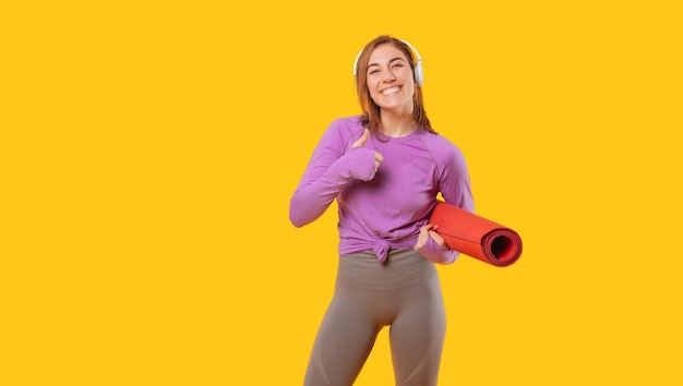 Веселая молодая женщина, держащая коврик для йоги, показывает большой палец вверх