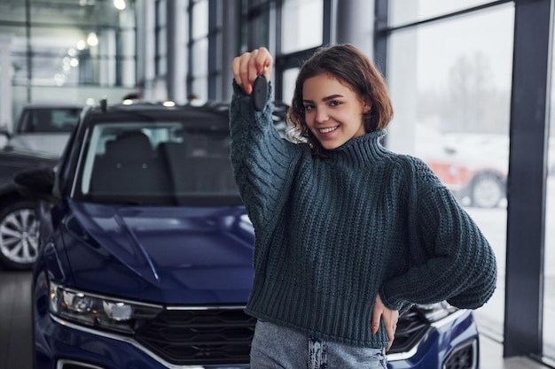 Cheerful young woman holds keys and standing in front of modern new car indoors.