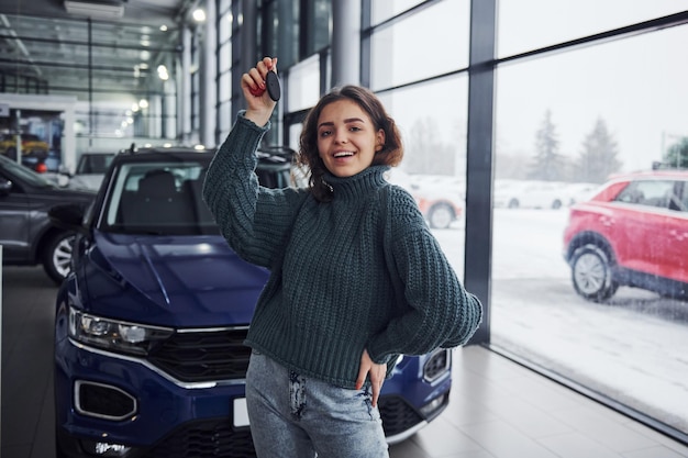 Photo cheerful young woman holds keys and standing in front of modern new car indoors.