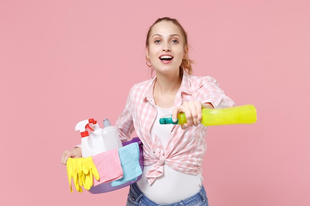 Cheerful young woman housewife in checkered shirt hold basin with detergent bottles, spray with washing cleansers while doing housework isolated on pastel pink background studio. Housekeeping concept.