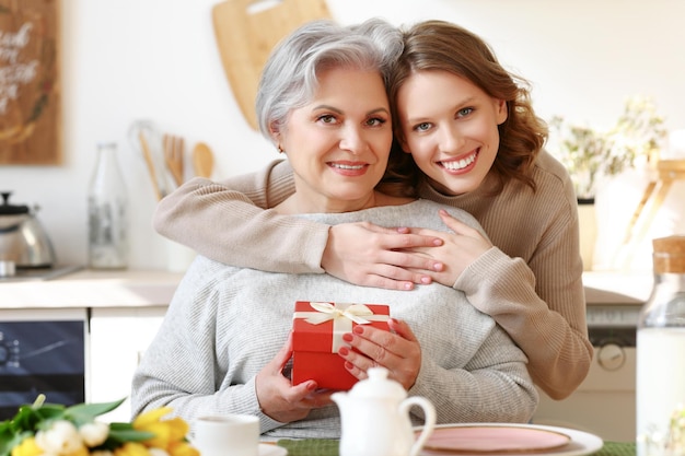 Photo cheerful young woman hugging happy elderly female and giving flowers and present box on mother day