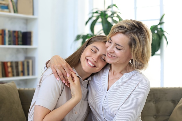 Cheerful young woman is embracing her middle aged mother in living room.