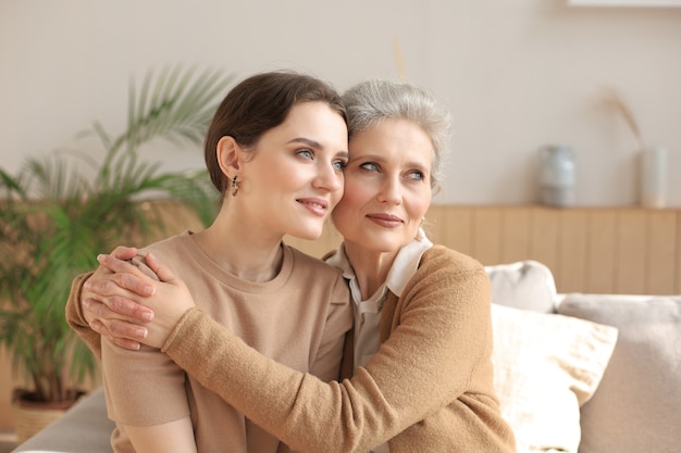 Cheerful young woman is embracing her middle aged mother looking away, touching cheeks, sitting on couch at home. Happy trusted relations. Family concept.