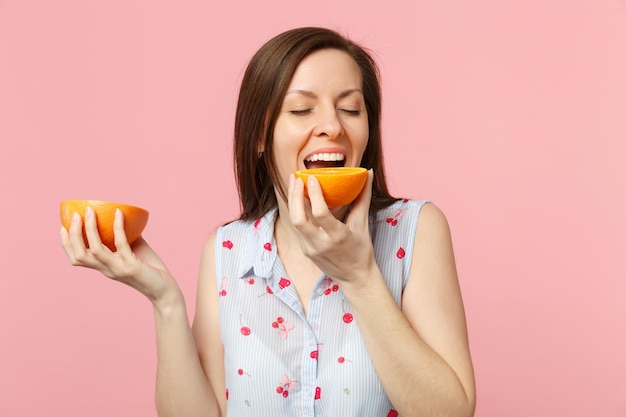 Cheerful young woman keeping eyes closed hold, biting halfs of fresh ripe grapefruit orange fruit isolated on pink pastel background. People vivid lifestyle relax vacation concept. Mock up copy space.