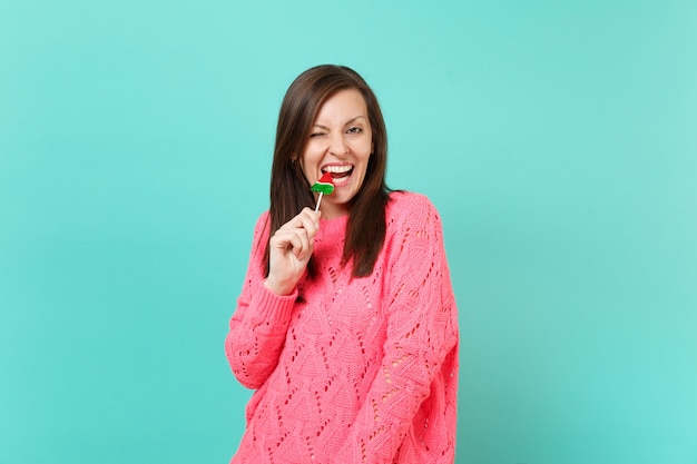 Cheerful young woman in knitted pink sweater blinking hold in hand biting watermelon lollipop isolated on blue turquoise wall background, studio portrait. People lifestyle concept. Mock up copy space.