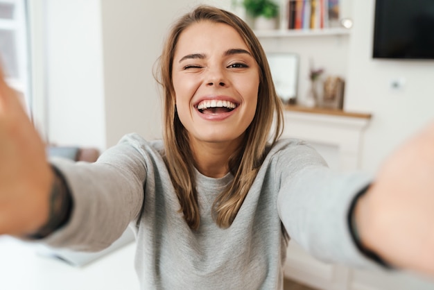 cheerful young woman laughing and winking while taking selfie at living room