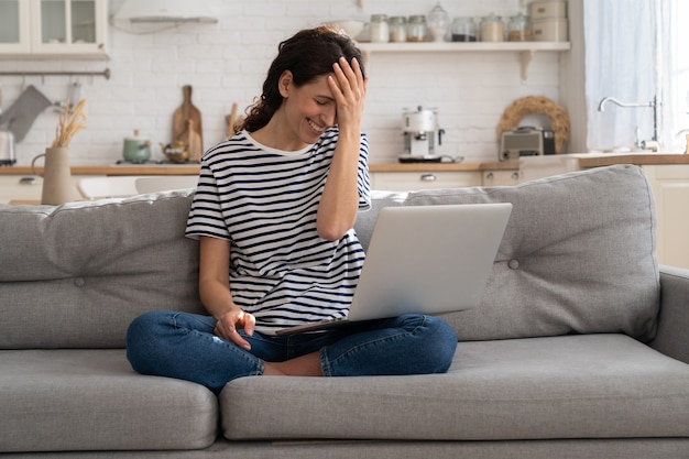 Cheerful young woman laughs while watches funny video on laptop at home