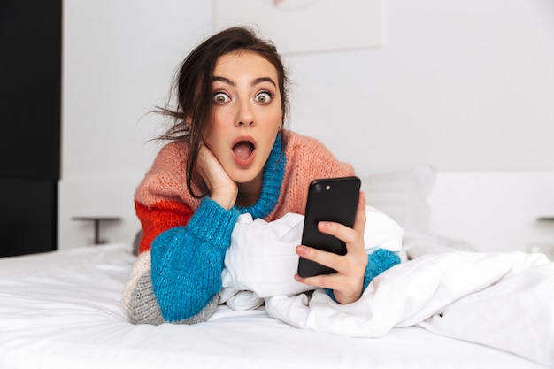 cheerful young woman laying on a bed at home, holding mobile phone