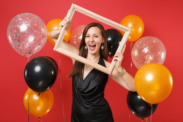 Cheerful young woman in little black dress celebrating and holding picture frame on bright red background air balloons. International Women's Day, Happy New Year birthday mockup holiday party concept.