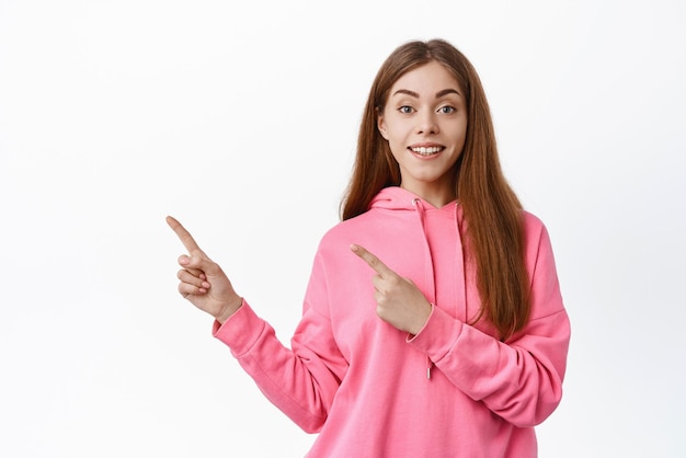 Cheerful young woman look with amazement and joy smile and point left showing copyspace aside standing over white background