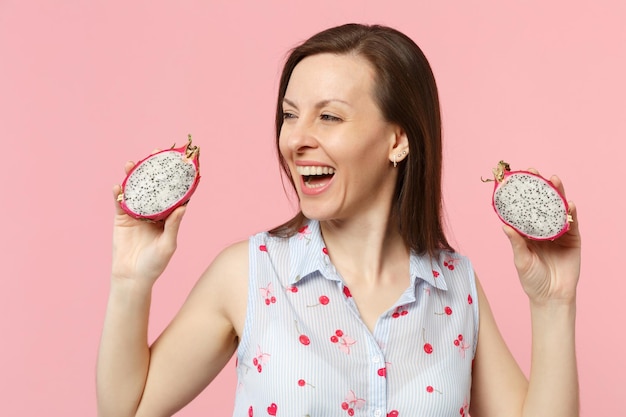 Cheerful young woman looking aside, holding halfs of fresh ripe pitahaya, dragon fruit isolated on pink pastel background in studio. People vivid lifestyle relax vacation concept. Mock up copy space.