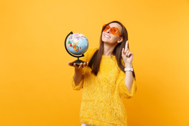 Cheerful young woman in orange heart eyeglasses dreaming about travel, pointing index finger up holding world globe isolated on yellow background. People sincere emotions, lifestyle. Advertising area.