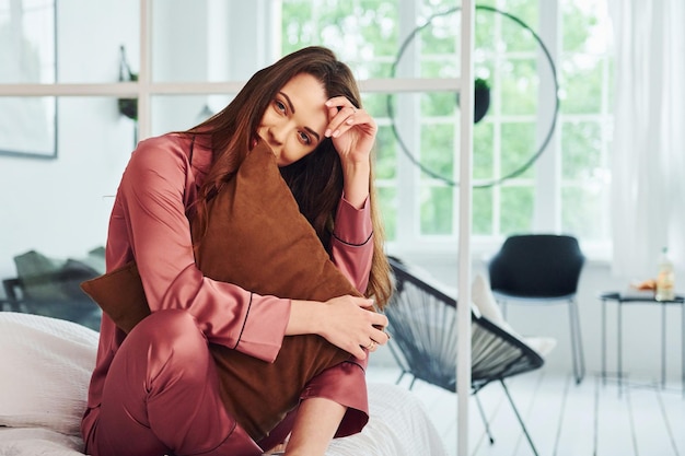 Cheerful young woman in pajamas sitting indoors at daytime and embracing the pillow