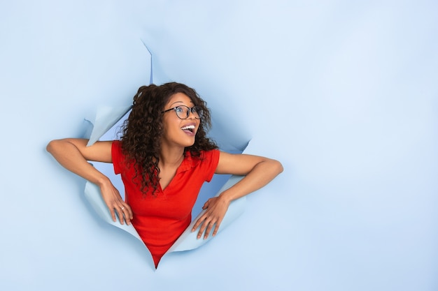 Cheerful young woman poses in torn blue paper hole background, emotional and expressive