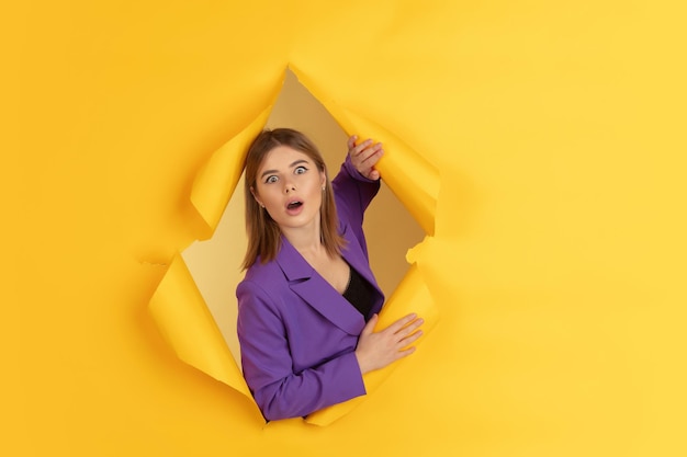 Cheerful young woman poses in torn yellow paper hole background, emotional and expressive