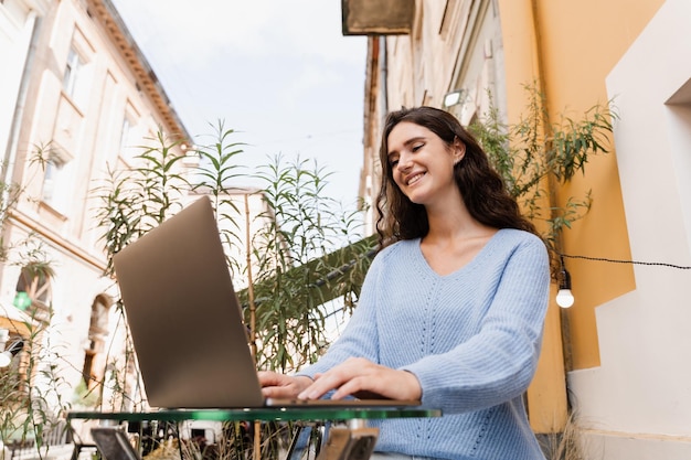 Cheerful young woman programmer works remotely on laptop and\
try to meet deadline outdoor in cafe candid girl with laptop is\
smiling and rejoices at successful work in it company