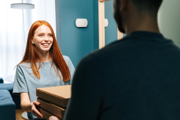 Cheerful young woman receiving paper boxes with hot pizza from unrecognizable delivery man on doorstep at home